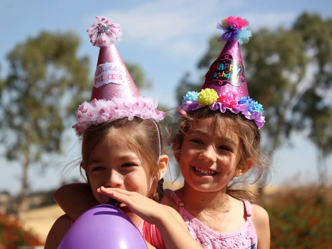 Zwei Kinder mit bunten Partyhüten feiern einen Kindergeburtstag im Freien